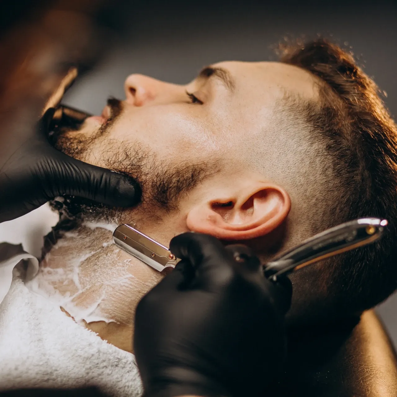 Barbero con guantes negros afeitando el cuello de un cliente con navaja sobre espuma de afeitar, con una toalla blanca sobre los hombros