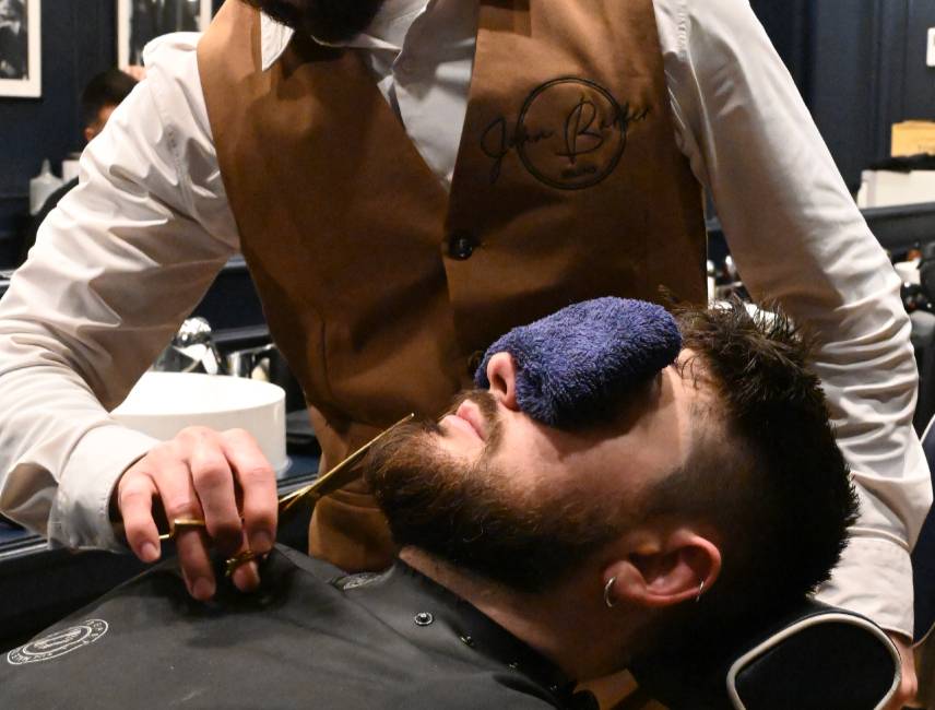 John-Barber-Barbier stutzt den Bart mit der Schere, ein Tuch über den Augen des Kunden: Schnitt und Rasur