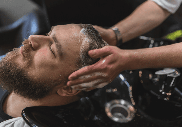 Homme barbu au bac pendant que le barbier lui masse le shampooing : shampooing et brushing chez John Barber