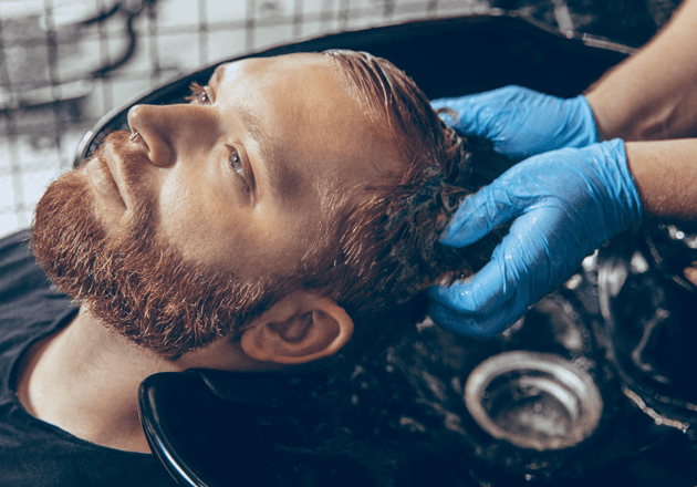 Mann mit kupferfarbenem Bart am Waschbecken, Hände in Handschuhen im Haar: stärkende Haarbehandlung