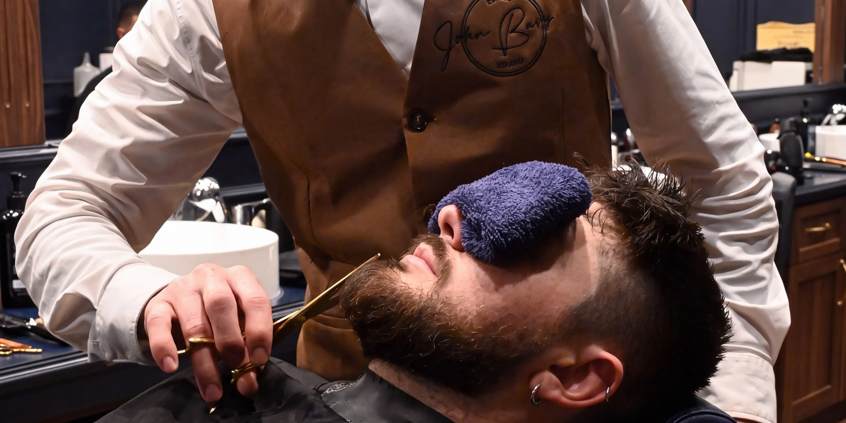 Barbier in Lederweste von John Barber stutzt den Bart eines Kunden mit der Schere, der Kunde hat ein warmes Handtuch auf den Augen